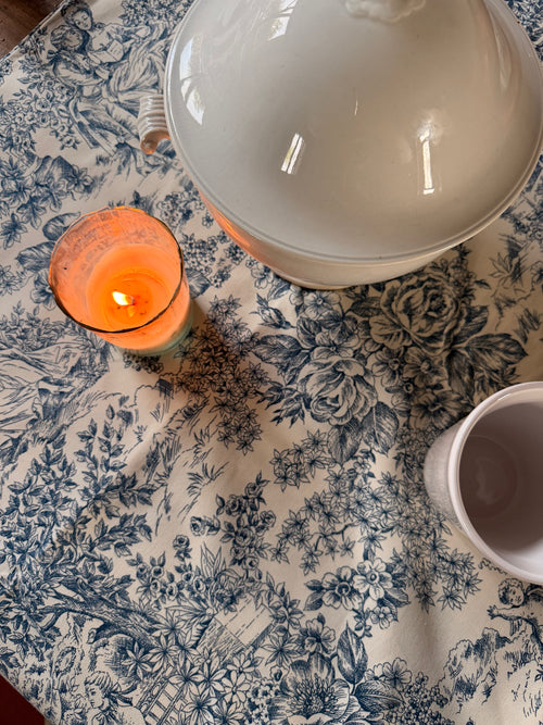 Nappe Louise à motif toile de Jouy bleu sur fond écru, ornée de scènes champêtres et de fleurs, présentée avec de la vaisselle ancienne – Maison Bellie.